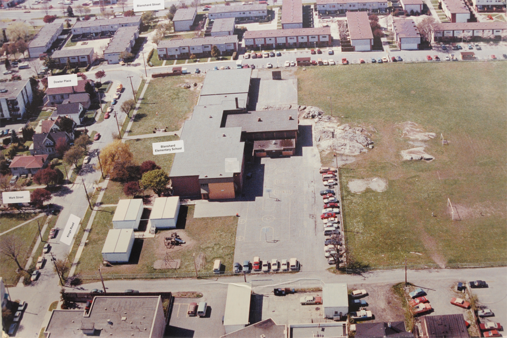 An aerial view of a school and playground, with a field on the right and the building on the left, surrounded by rows of homes and other structures