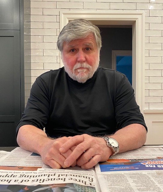 A man with light grey hair and a short beard sits with his hands clasped together in front of him on a table covered with newspaper
