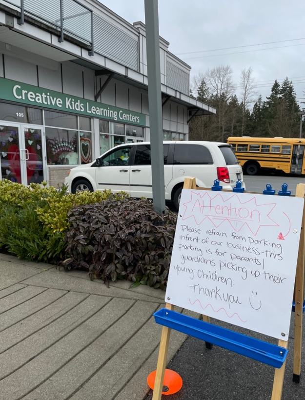 A sign asking protesters not to park in front of a business is set up in front of a daycare parking lot, where a white mini van and a school bus are parked