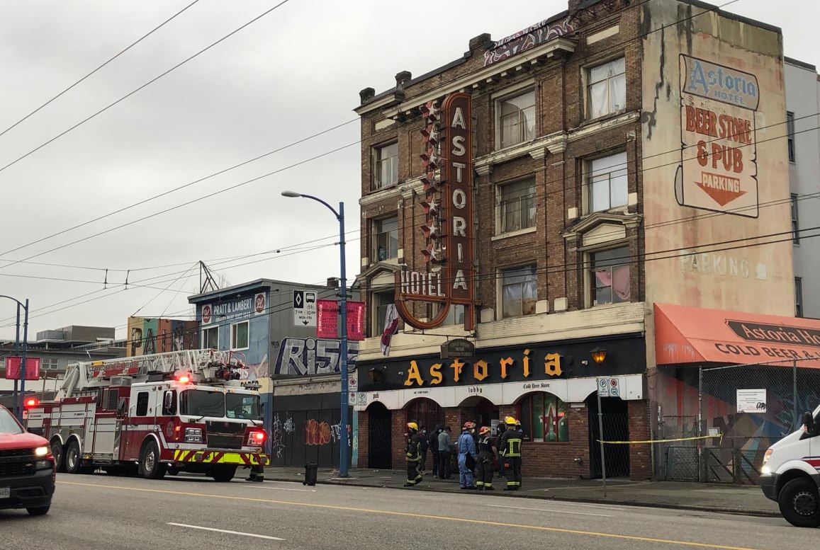 A brown brick building with Astoria written on a vertical sign as well as in yellow on a black awning is seen in front of a light grey sky. THere' a fire truck parked in the lower left corner of the frame, and police tape around the building