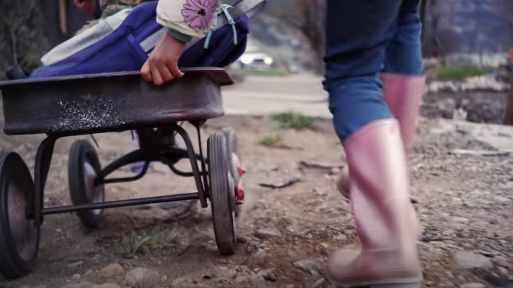 A child's pink boots are seen walking through dirt, while a girl's hand pushes a wagon