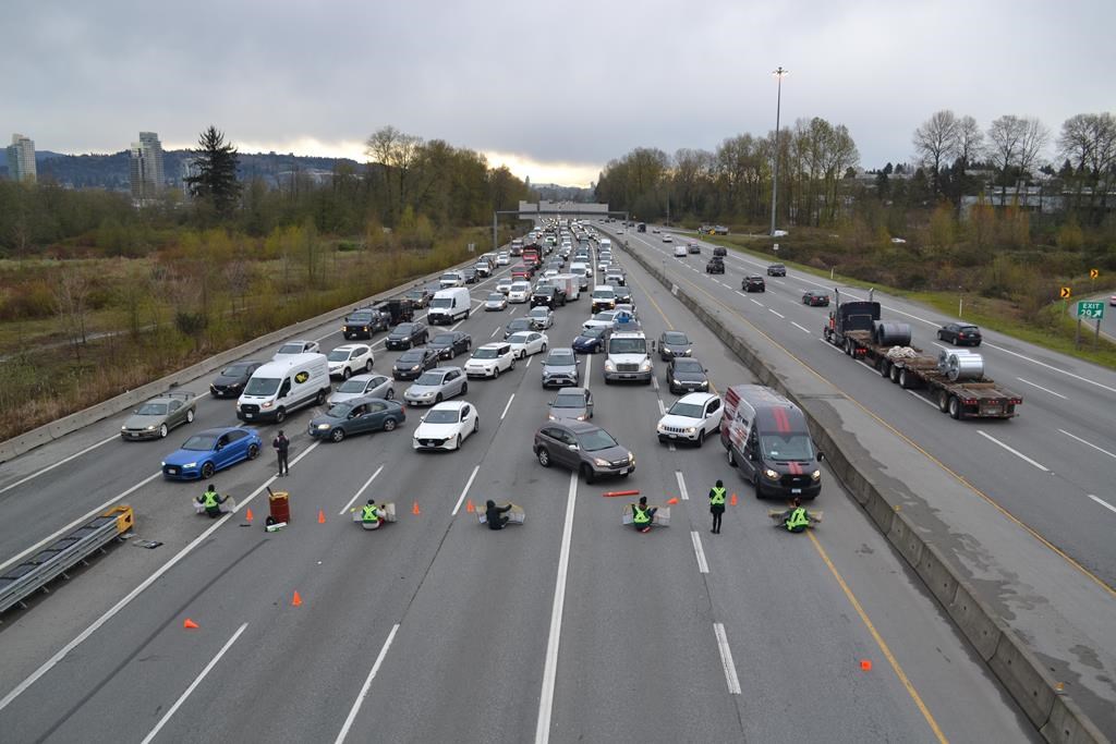 A row of people sits across a busy highway, with a lineup of vehicles blocked by the demonstrration