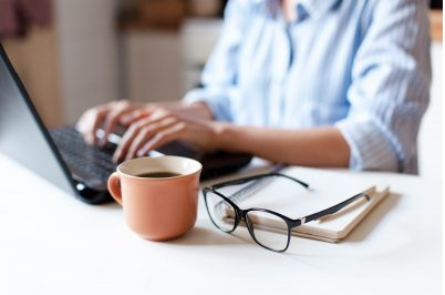 A photo of a person on a computer with a cup of coffee and their glasses and a note pad.