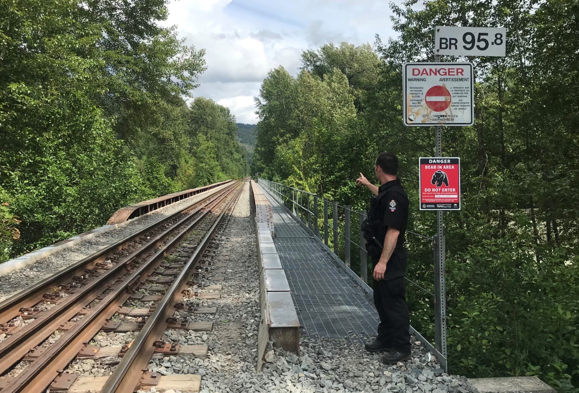 An officer stands at the beginning of a rail bridge pointing away from the camera, standing next to a sign warning of bears in the area