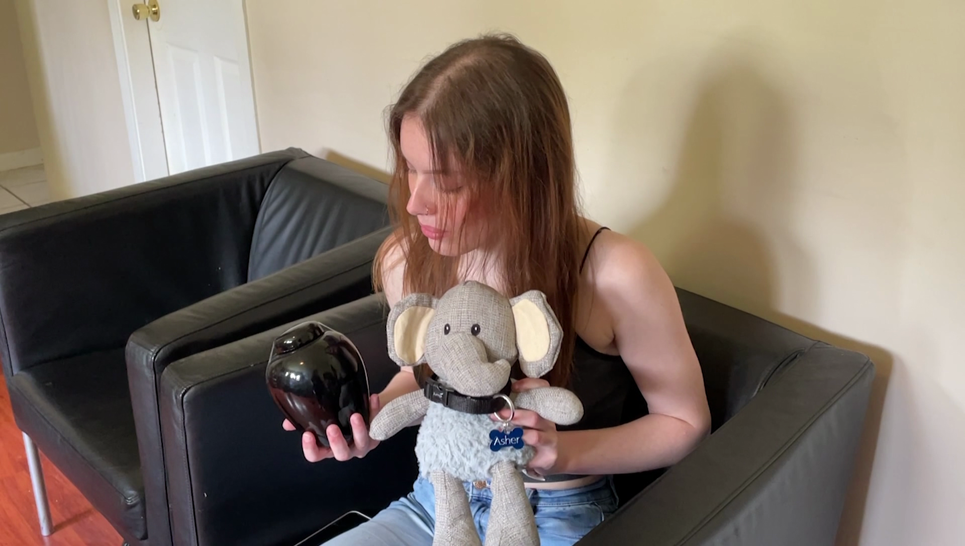 A woman who is sitting in a black armchair holds up a black urn in one hand and an elephant stuffed animal in the left, the elephant with a coller and tag that reads Asher on it