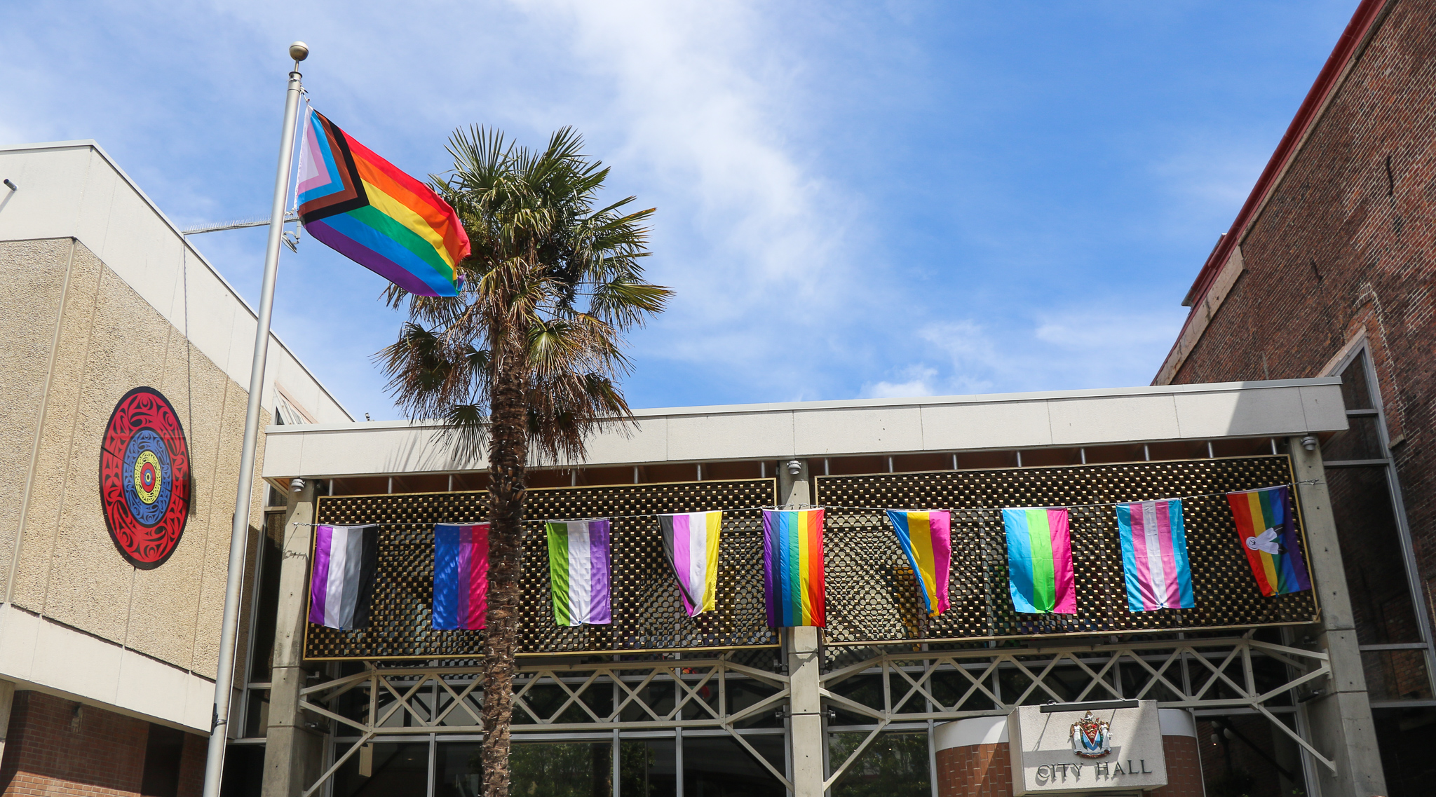 A Pride flag on top of a flag pole waves in the wind outside Victoria City Hall, where other smaller Pride flags are hung up on the building