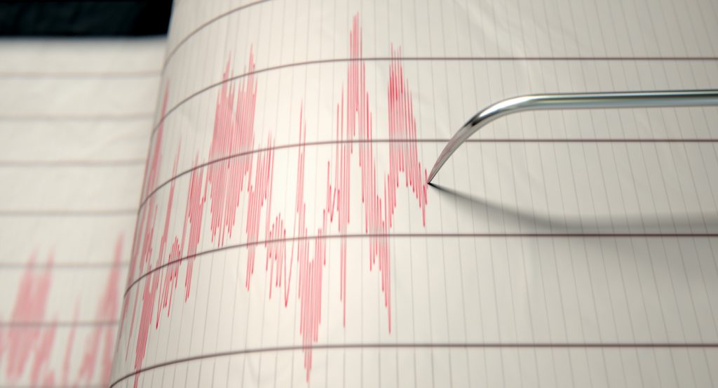A closeup of a seismograph machine needle drawing a red line on graph paper depicting seismic and earthquake activity