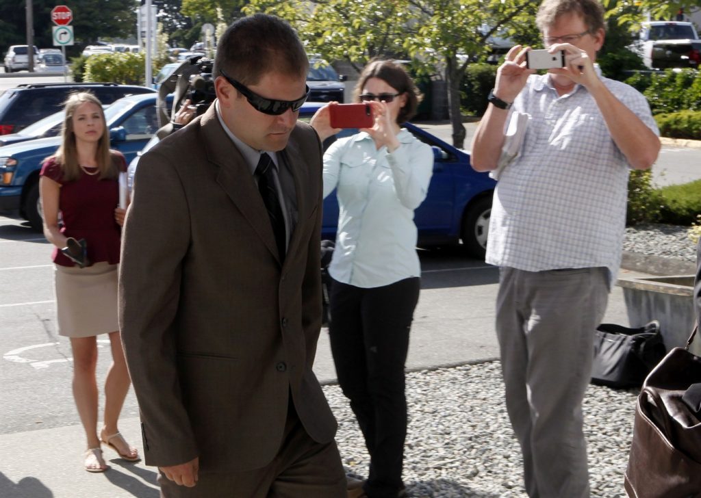 A man wearing a brown suit and sunglasses walks with his head down as three people behind him in the background take photos of him with their cellphones