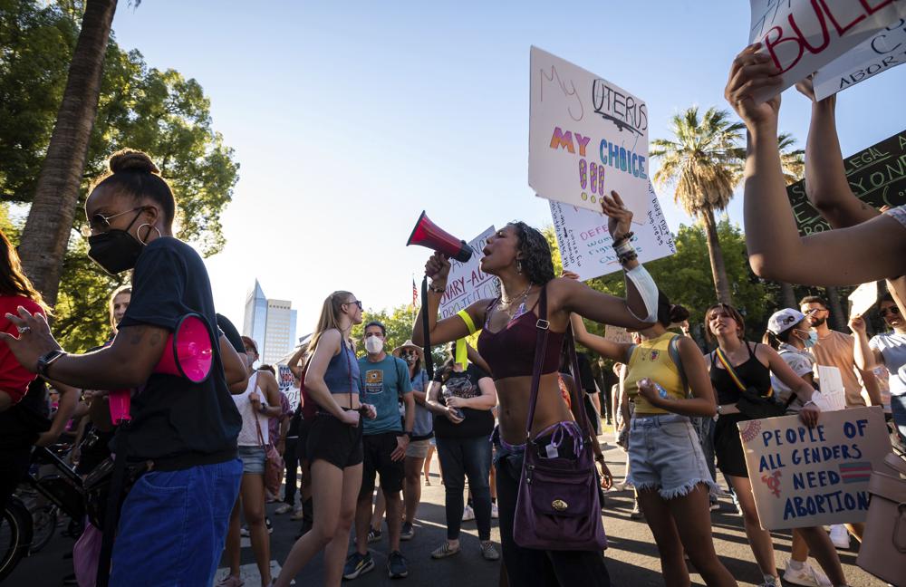 A woman speaks into a bullhorn while carrying a sign in the other hand, surrounded by protesters