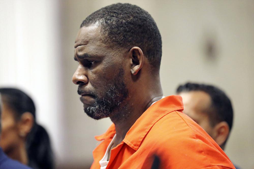 R. Kelly stands facing the left of the screen wearing an orange prison jumpsuit