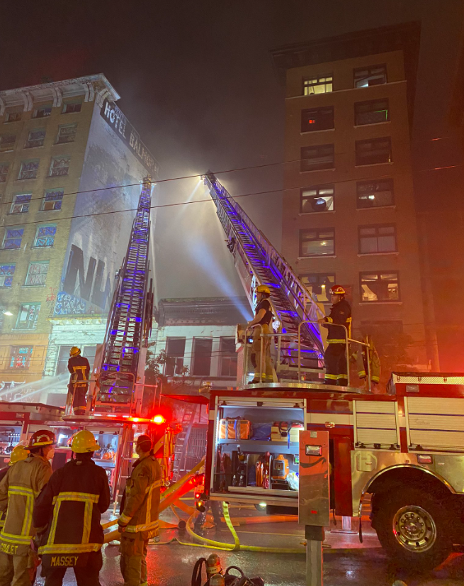 Firefighters stand next to two fire trucks with their ladders up toward a building in the early morning as they battle flames at a building