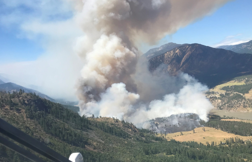 An aerial view of a wildfire burning through trees and fields in B.C., with mountains in the background and foreground