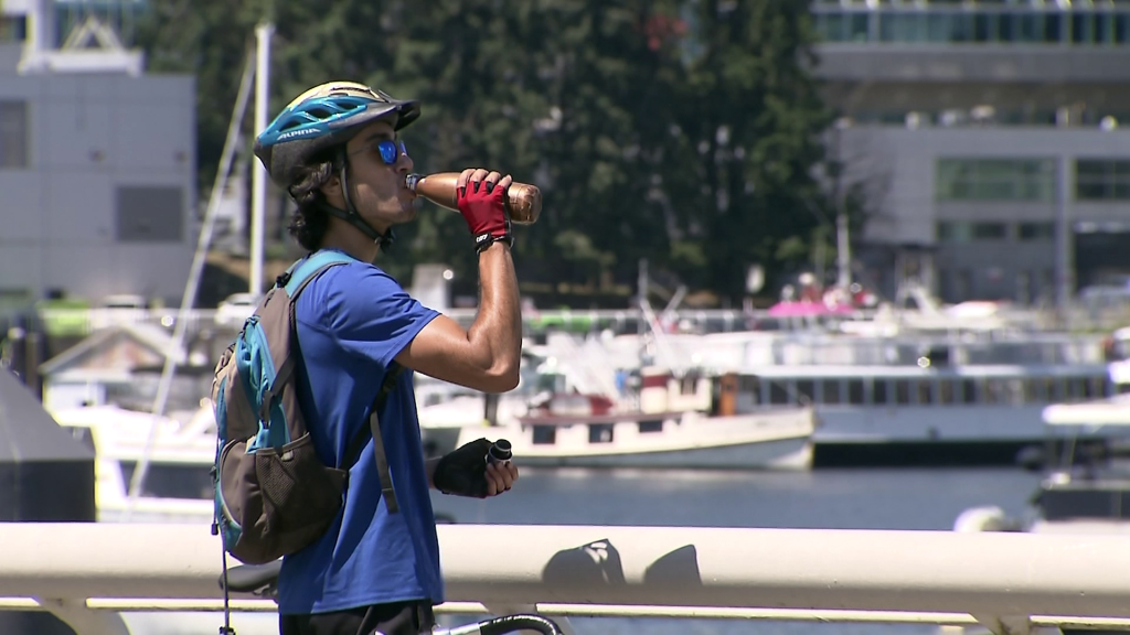 A person wearing a bike helmet and sunglasses drinks from a reusable water bottle in front of a railing by the water, boats in the background