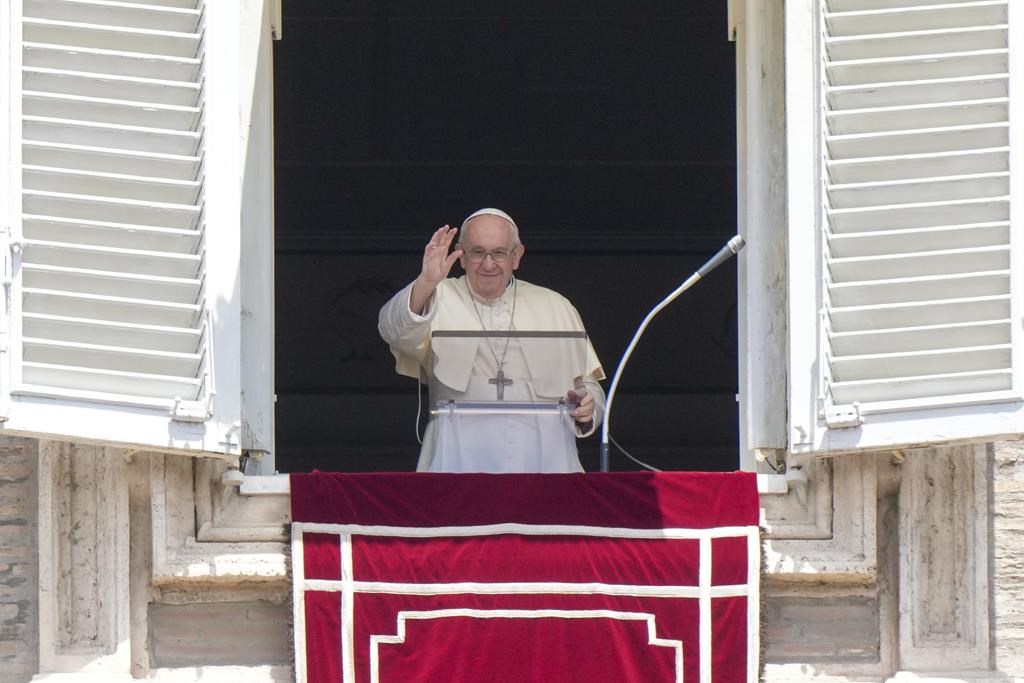 The Pope recites a prayer from a window overlooking St. Peter's Square, at the Vatican. He has his right hand raised up and is smiling