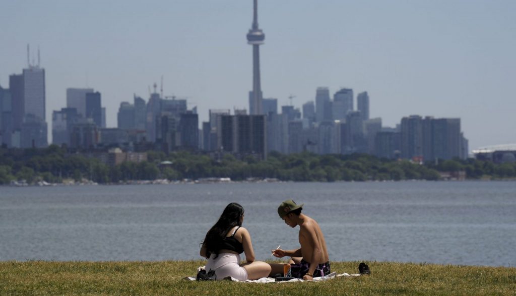 Two people sit outside on some grass with water and the Toronto skyline in the background on a summer's day