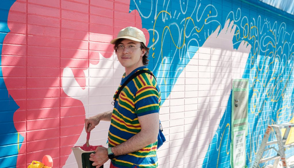 A man wearing a stripped shirt and a ballcap stands in front of a mural with a bucket of paint and a paintbrush as he looks left toward the camera