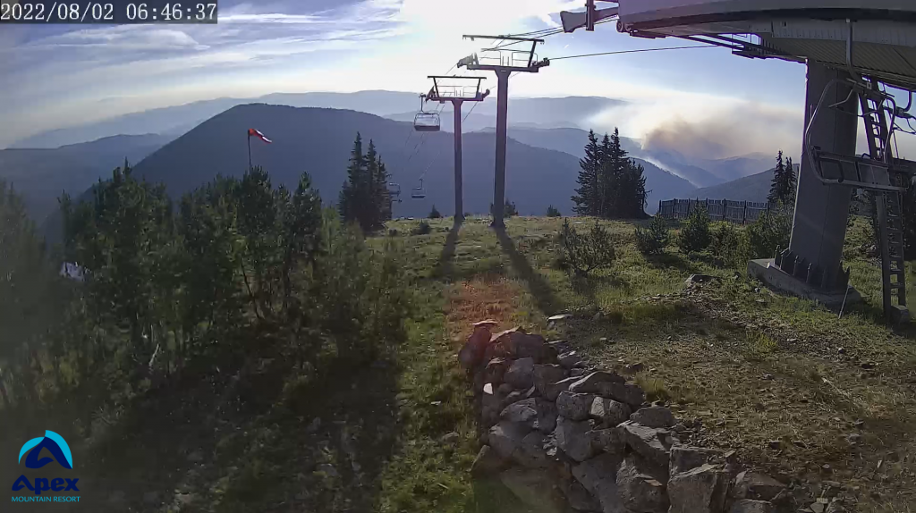 A webcam shot from the summer months when there is no snow shows a mountain range in the distance with smoke from a wildfire along one section. The photo also shows a chair lift in the foreground