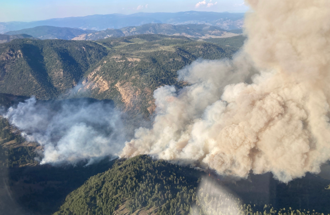 Thick smoke from a wildfire rises from a forested mountain ridge. The fire is pictured from above