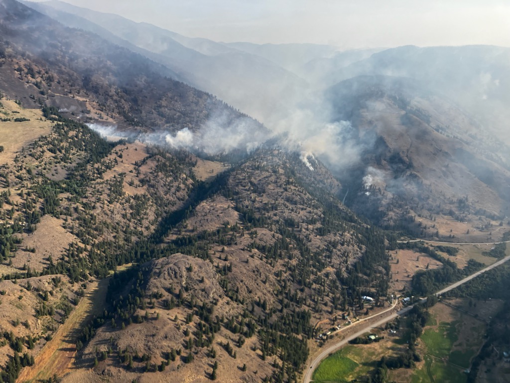 Thick smoke from a wildfire rises from a forested mountain ridge. The fire is pictured from above