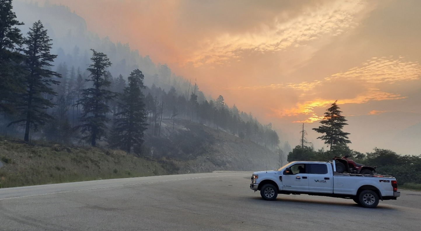 A BC Wildfire Service pick up truck is parked at the edge of a highway in front of a forested mountain side where smoke is rising from wildfires. The sun is seen rising through some clouds and smoke in the distance