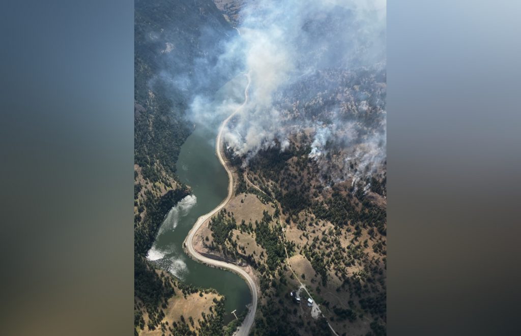 This aerial photo shows smoke rising from a wildfire burning in forested areas near a river and a winding highway road