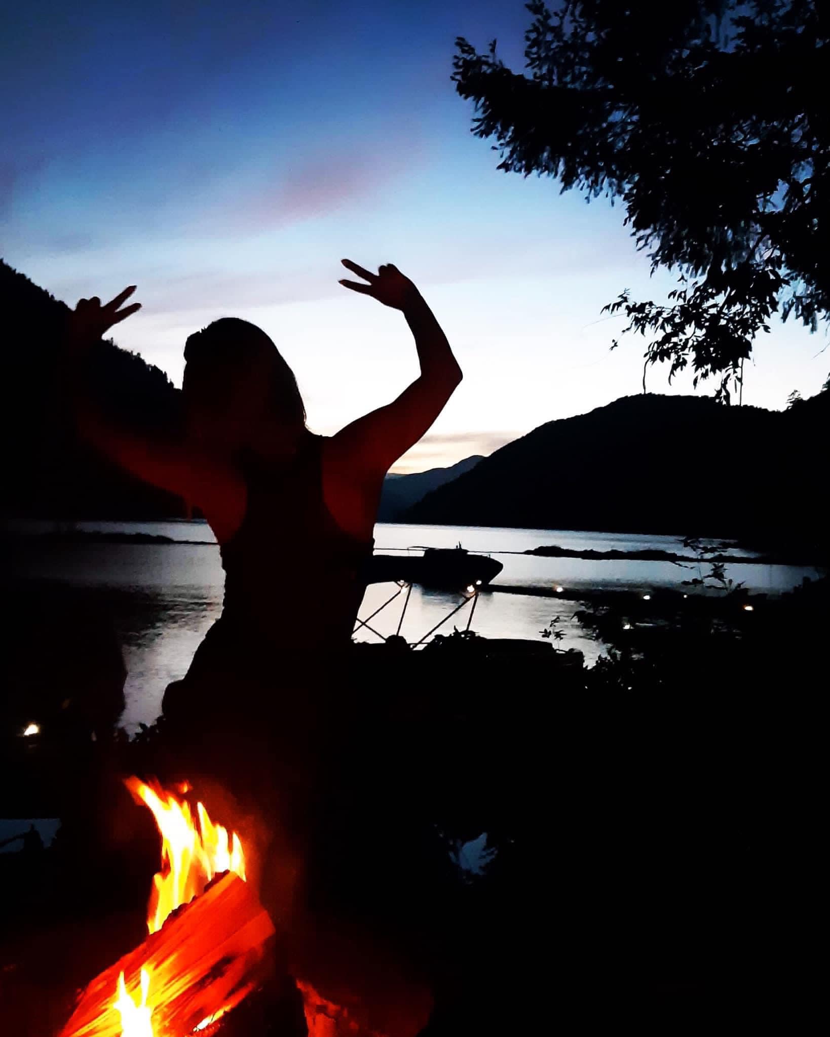 A silhouette of a woman holding up her arms behind a campfire with a lake and darkening sky in the background