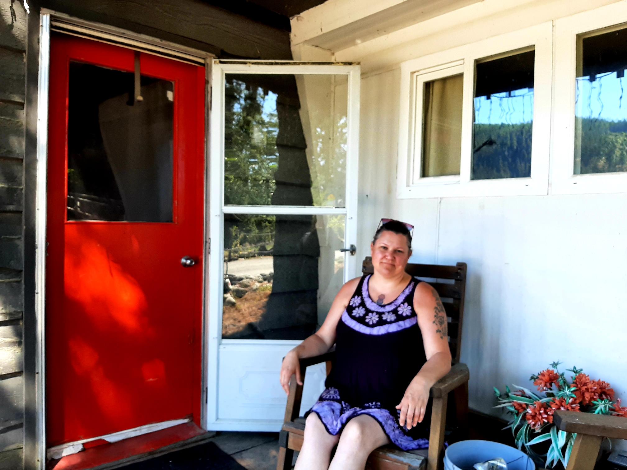 A woman sits on a chair outside a home on the porch. There's a red door to her right and a white trim behind her