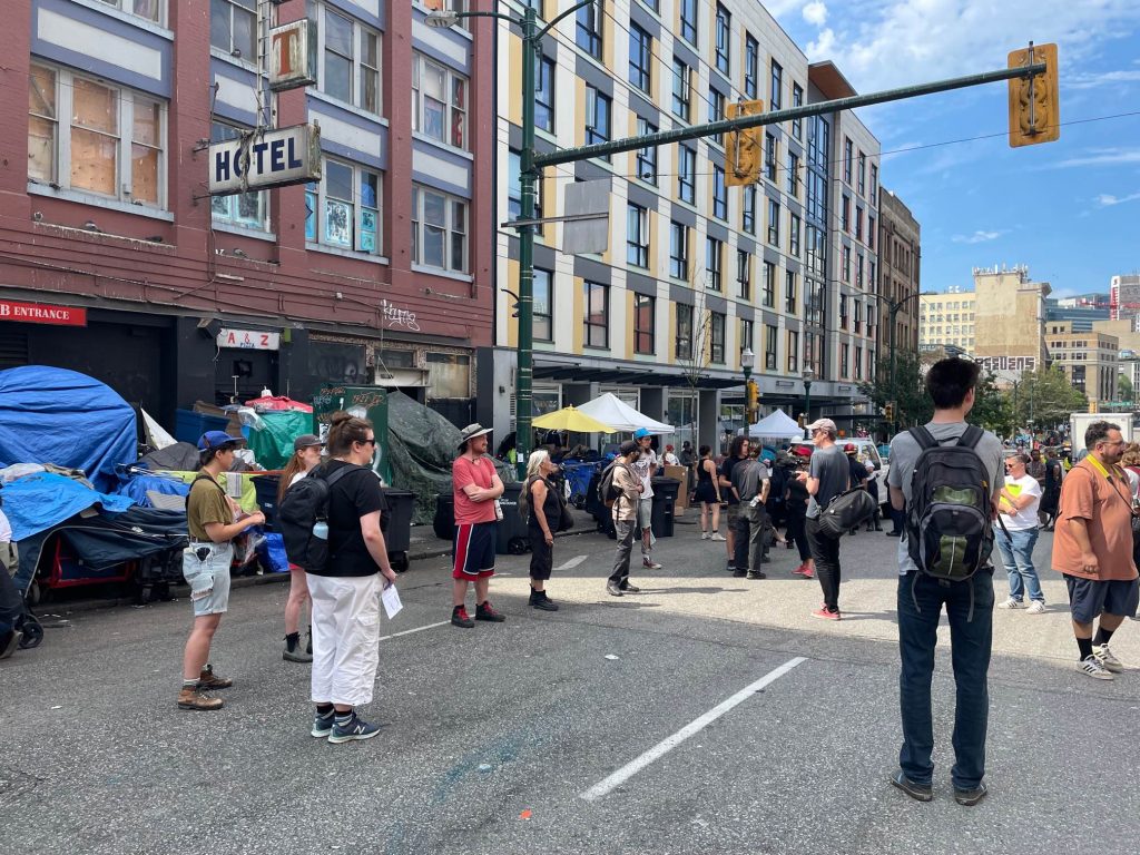 People stand in the middle of a blocked-off road as tents and other belongings are cleared from a sidewalk