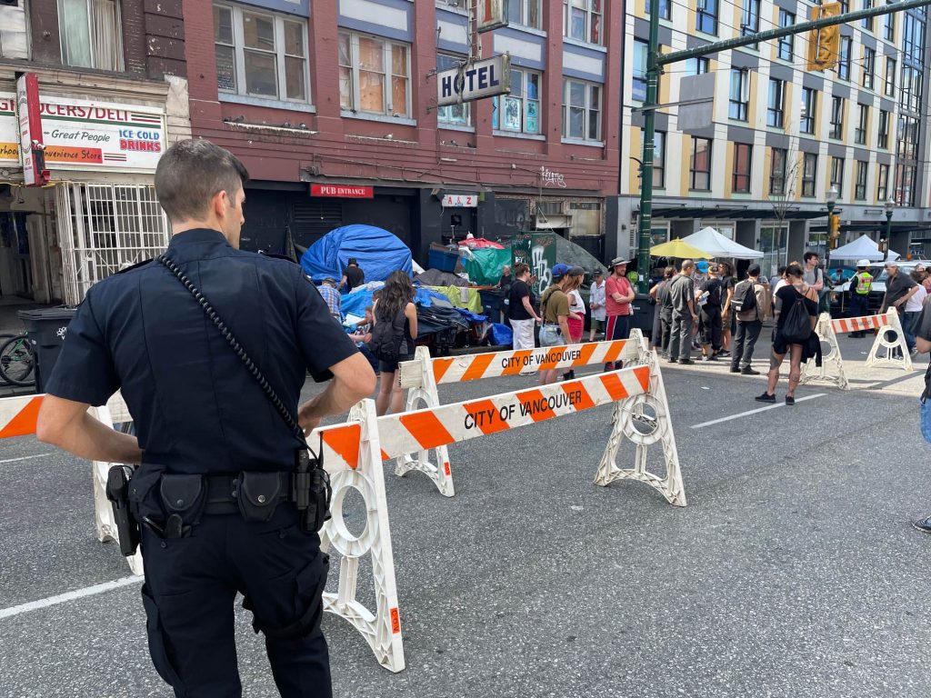 A police officer looks on behind a City of Vancouver barricade as people remove tents and other belongings from a sidewalk