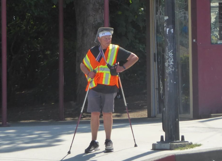 A 79-year-old man stands at a street corner with two walking poles while wearing a high-visibility vest, a head band, and hat