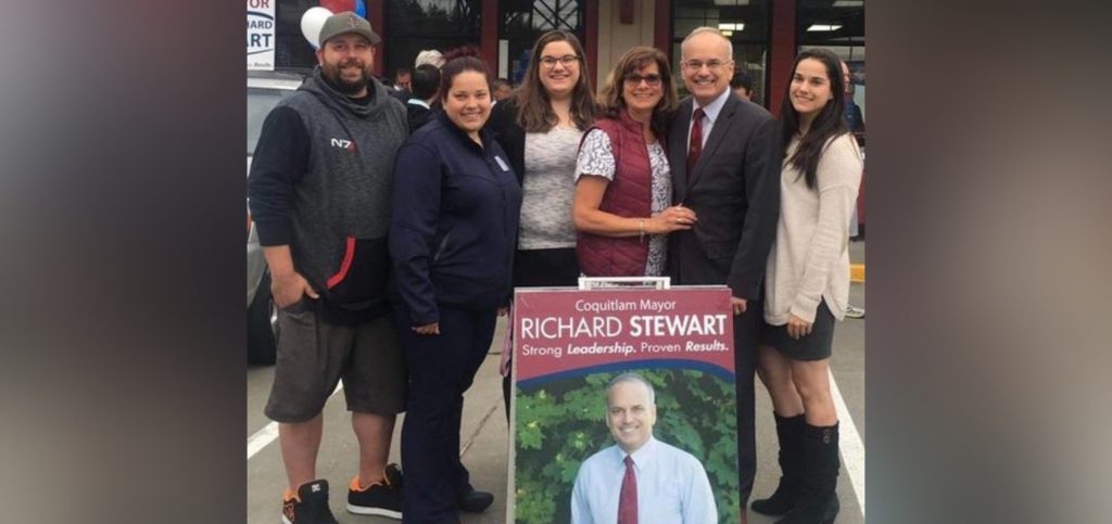 A group of six people stand side by side, posing for a photo, behind an election sign for Richard Stewart