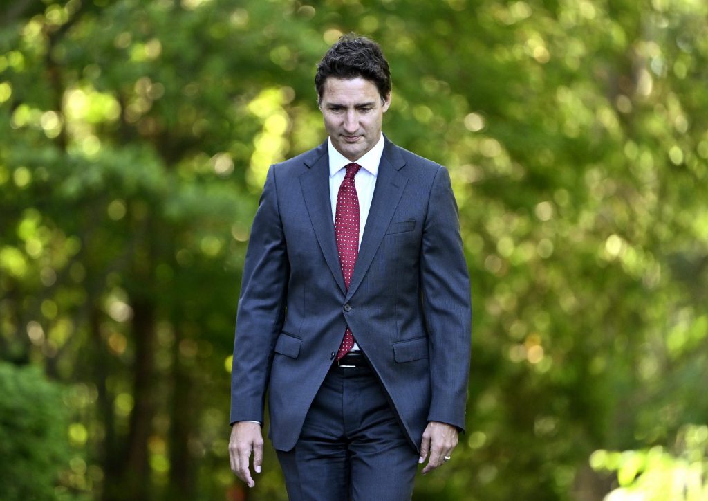 Prime Minister Justin Trudeau wearing a dark blue suit and a red tie walks along a path with trees in the background