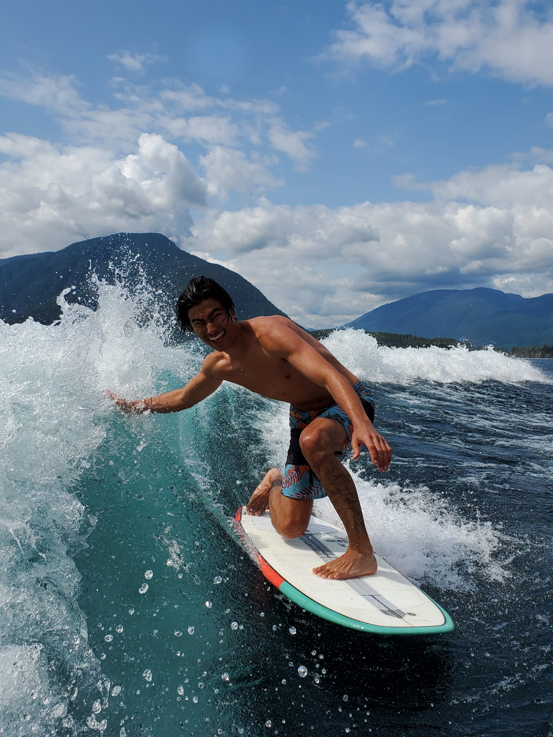 Jason Ono-O'Connor - Barrie crash 2 A man rides a surf board on a wake in the middle of an open body of water, with mountains in the background