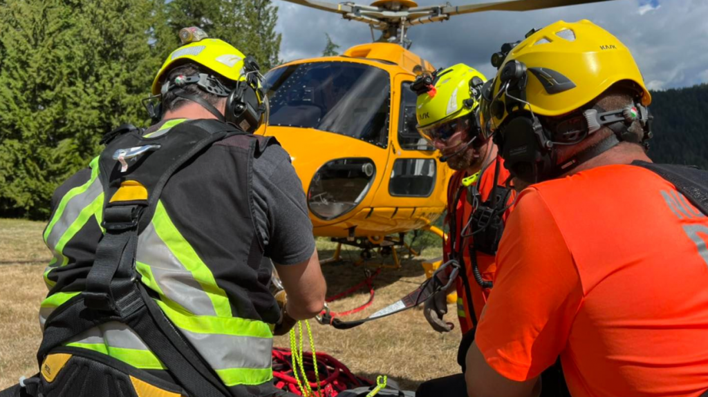 Three search and rescue volunteers wearing high-viz clothing and helmets are huddled in a group with a helicopter in the background