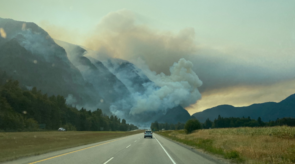 A fire along a mountainside in the distance sends thick smoke into the sky. A road with a car in the distance is pictured in the foreground