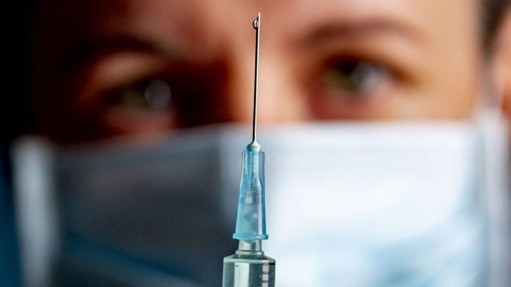 A vaccine is prepared as a person wearing a mask looks on from behind it