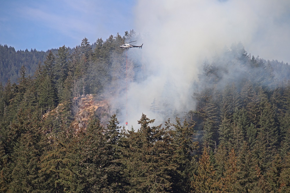 Smoke rises from a forested area as a helicopter drops water on a wildfire