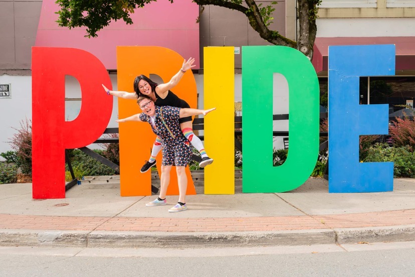 Teri Westerby and Mallory Tomlinson posing in front of a rainbow coloured Pride sign