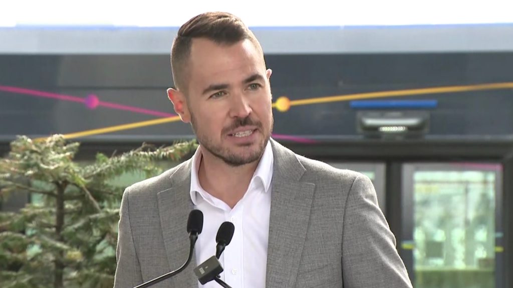 Port Coquitlam Mayor Brad West speaks at a podium with a transit bus in the background