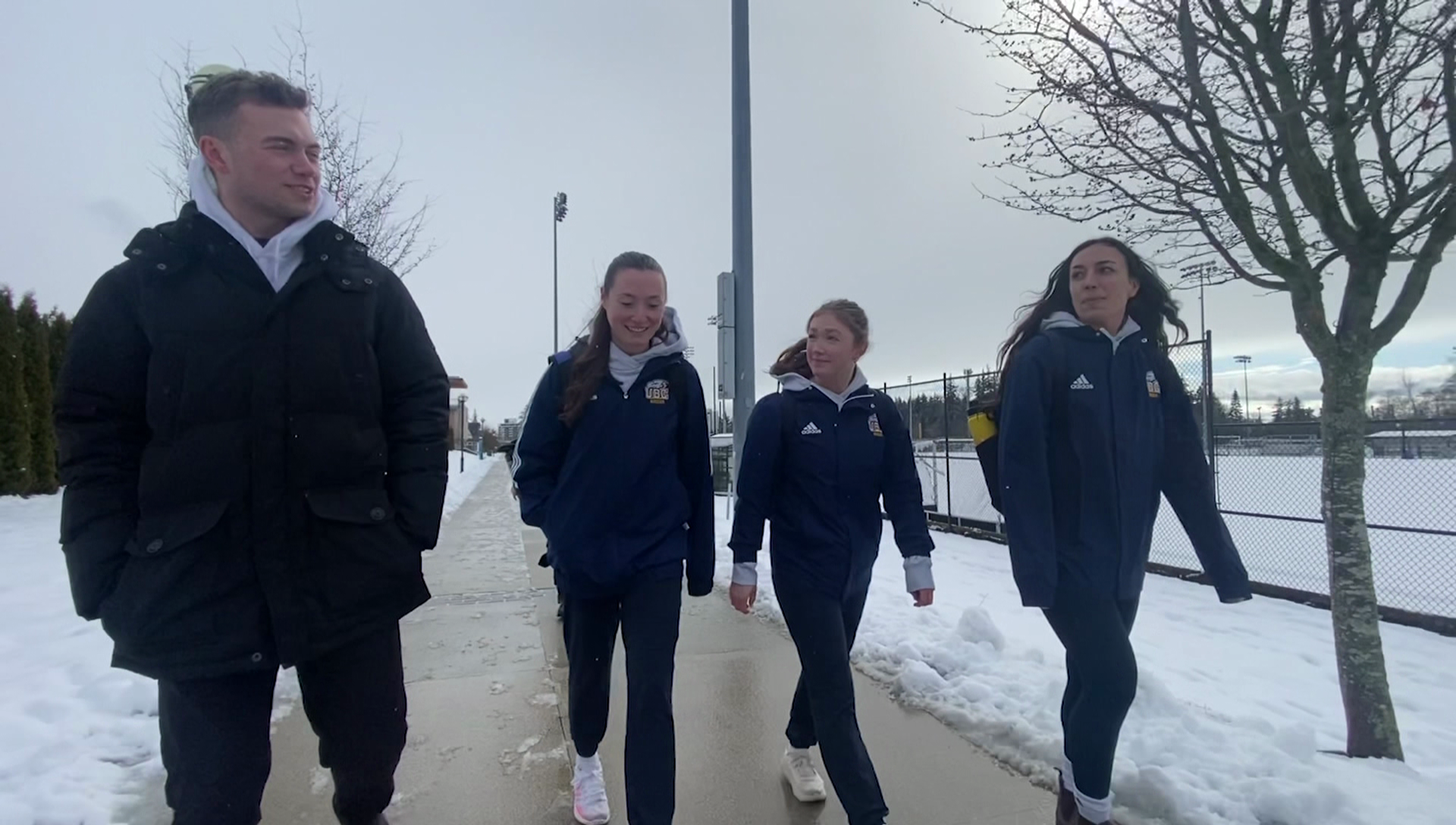 Soccer players walk on a sidewalk past a snowy field.