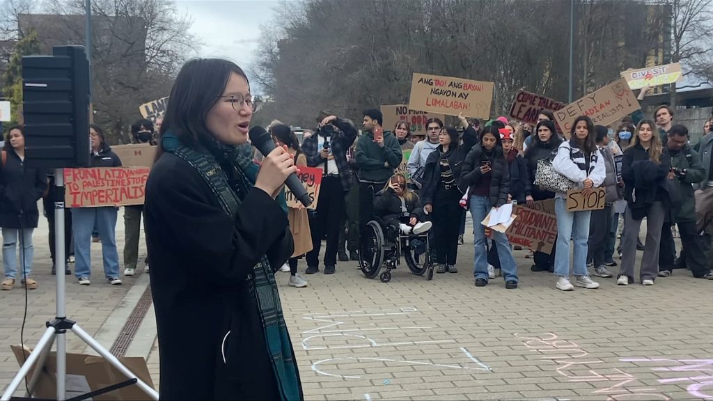 students gathered at the university of british columbia to call for the school to make better investments.. a woman stands in front of a group of people with a microphone.