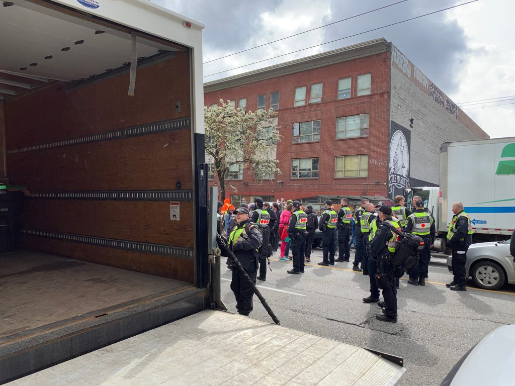 a group of police officers stands on the road near a large van. On Wednesady part of East Hastings street was shut down as part of the city's plan to decamp the area.