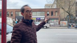 man wearing glasses and maroon jacekt points at buildingHenry Tsang is an author and historian that focuses on the anti asain riots of 1907 in Vancouver.