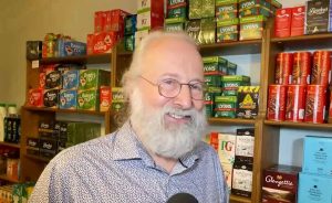 Stephen Coulson a man with a large white beard stands in front of shelving unit with cans and british goods.