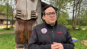 a man stands in a black jacked in front of a large bear carving. he is an elected member of the squamish nation and speaking on the return of Indigenous artifacts to B.C.