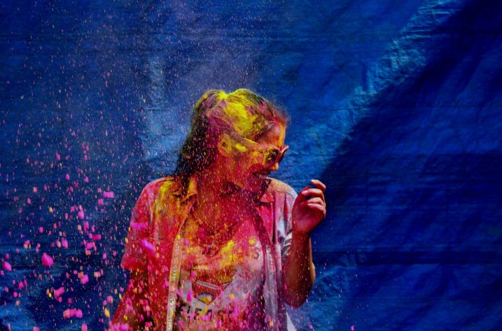 a woman stands in front of a blue tarp while people throw colourful paint-like powder at her in celebration of colourfest