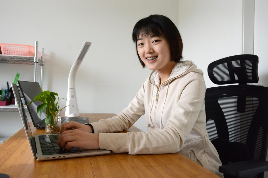 A young student sits at a table smiling in front of a laptop.