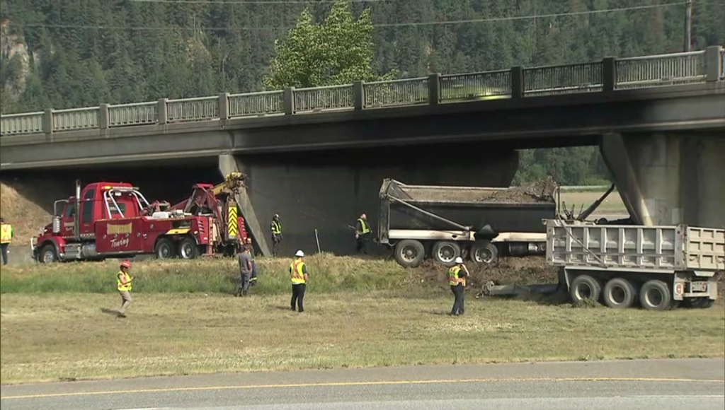 Crews stand in a field next to a dump truck that crashed into an overpass