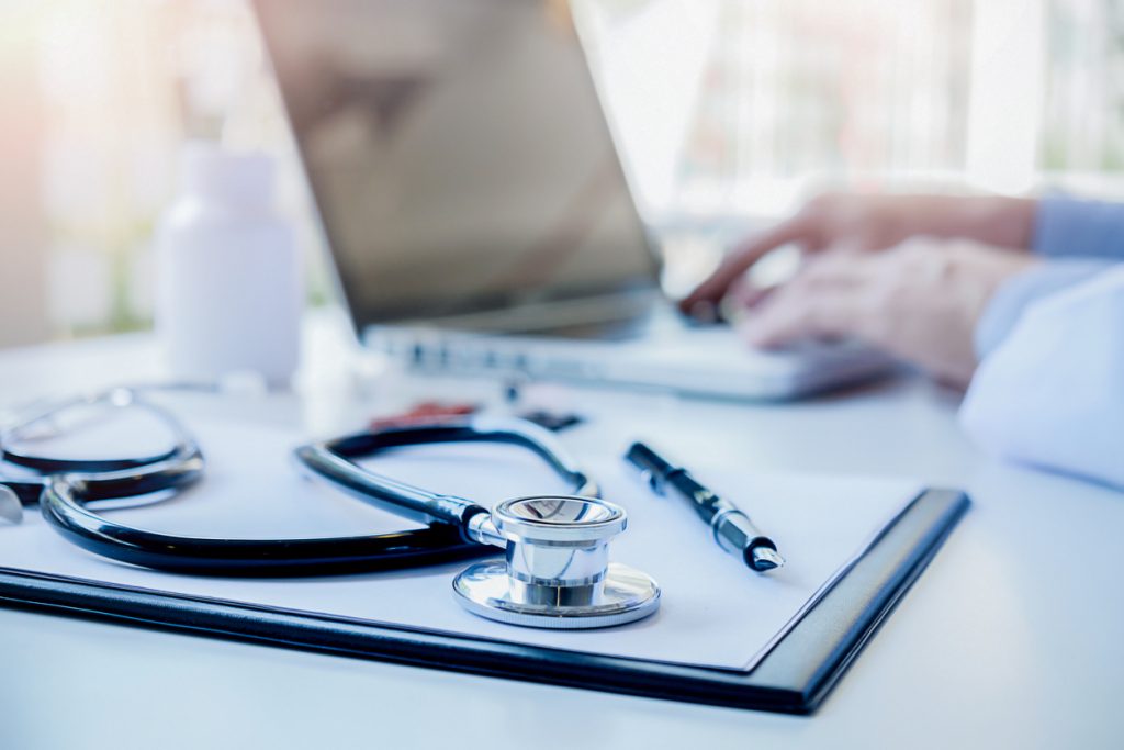 A stethoscope sits on the desk as a doctor types on a laptop