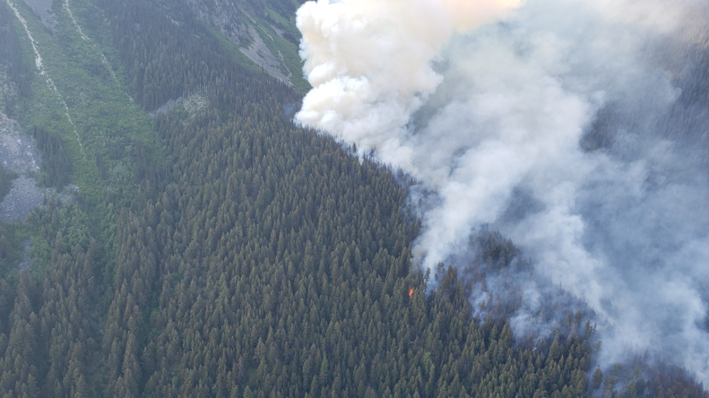 Smoke rises from flames on the side of heavily forested mountain. The Casper Creek wildfire is burning west of Lillooet in the vicinity of Anderson Lake